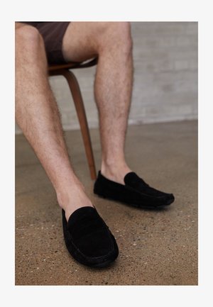 Mocassini in suede neri presenta un design in stile moccasin con dettagli cuciti, suole piatte e una forma slip-on comoda.