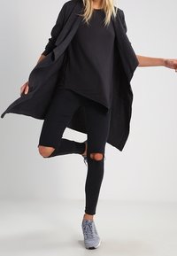 Czarny długi kardigan na czarnym topie, w połączeniu z przetartymi czarnymi dżinsami i szarymi trampkami. Skupienie na warstwowaniu i szczegółach zniszczonego denimu.