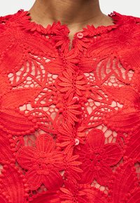 Κοντινό σε κόκκινο δαντελωτό ένδυμα με λουλουδάτα σχέδια και κλεισίματα με κουμπιά, φορεμένο στο πάνω μέρος του στήθους και του λαιμού.
