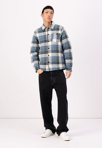 Quilted flannel jacket featuring a blue and black check pattern, lightweight construction, and front pocket, paired with black pants and white sneakers.