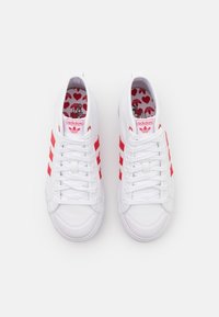 White sneakers with three red stripes on each side, featuring a textured design and a printed insole with red hearts.