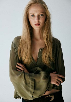 Mujer joven con cabello largo y rubio que lleva una blusa verde oliva con mangas anchas, brazos cruzados, mirando directamente a la cámara.