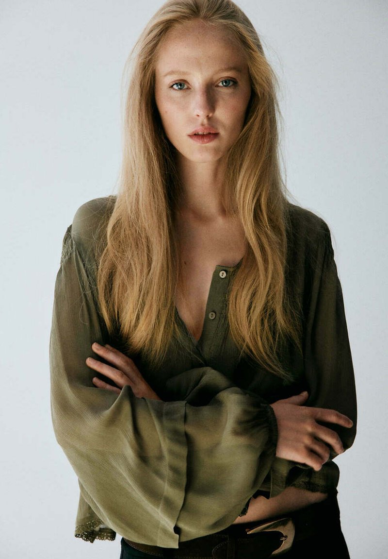 Jeune femme aux longs cheveux blonds portant une blouse vert olive à larges manches, bras croisés, regardant droit dans la caméra.