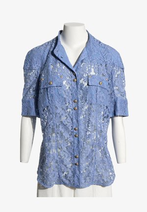 Blusa de encaje azul claro de manga corta con botones dorados y dos bolsillos en el pecho sobre un torso de maniquí blanco.