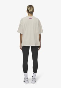 Beige Übergrößen-T-Shirt mit überschnittenen Schultern und einem pinken Logo-Detail auf dem Rücken, kombiniert mit schwarzen Leggings und weißen Turnschuhen.