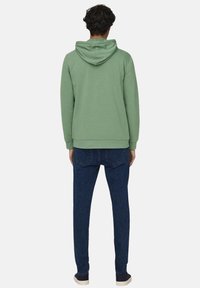 Sudadera verde con capucha y bolsillo tipo canguro, combinada con jeans ajustados de color azul oscuro y zapatillas negras. Tejido suave y corte relajado.