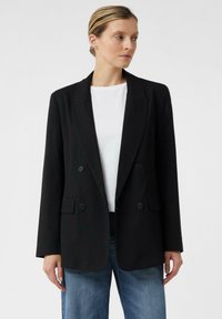 Donna che indossa un blazer nero doppiopetto sopra una camicia bianca e jeans blu, in piedi di fronte a uno sfondo chiaro e uniforme.
