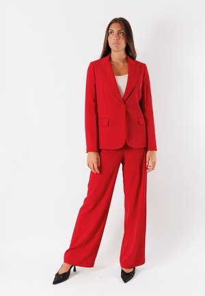 Blazer rosso su misura con un solo bottone e tasche a filetto, abbinato a pantaloni larghi coordinati. Tacchi neri a punta completano l'outfit.