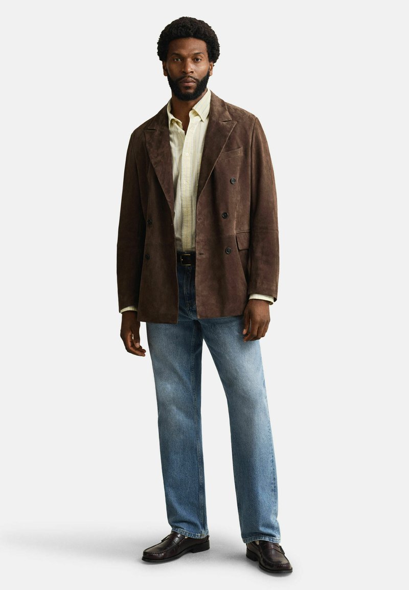 Hombre con chaqueta marrón de ante de doble botonadura, camisa de rayas amarillas claras, vaqueros azules y mocasines marrón oscuro, de pie sobre fondo blanco.