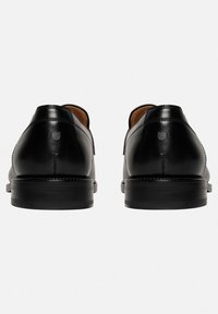 Chaussures en cuir noir avec une texture lisse, un talon arrondi et des détails de couture visibles. Design minimaliste avec un petit accent de logo.