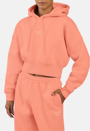 Vrouw draagt een zalmroze cropped hoodie van Reebok en bijpassende sweatpants, met haar hand in de zak, tegen een lichte achtergrond.