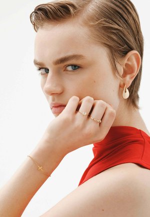 Las joyas de oro incluyen un pendiente en forma de gota torcida, un anillo brillante, una pulsera delicada con una cruz y una blusa roja ajustada.
