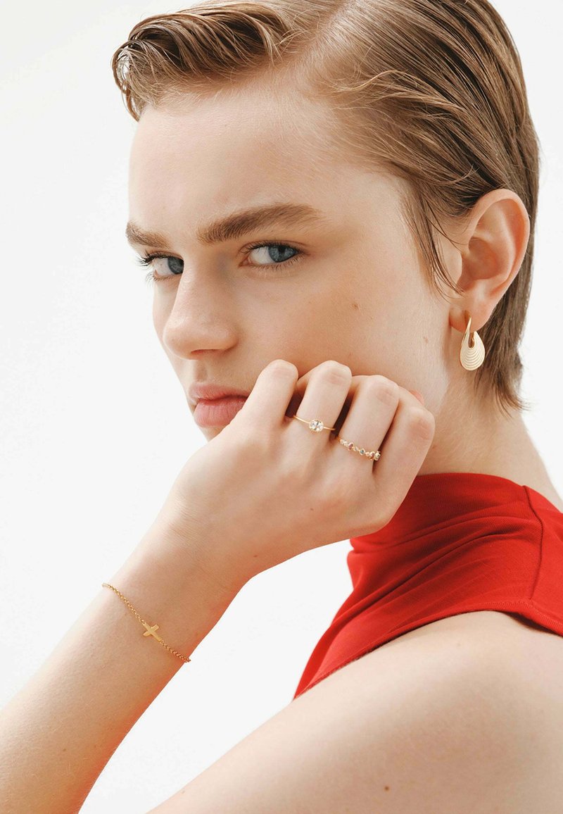 Las joyas de oro incluyen un pendiente en forma de gota torcida, un anillo brillante, una pulsera delicada con una cruz y una blusa roja ajustada.