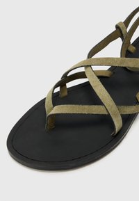 Sandali in suede verde oliva con cinturini incrociati, una suola nera liscia e un design minimalista, caratterizzati da un profilo piatto e senza decorazioni.