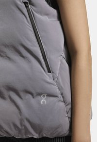 Lys grå puffer vest med en blank overflade, der har en sidelukning og et trykt logo nederst til højre. Stoffet ser blødt og isoleret ud.