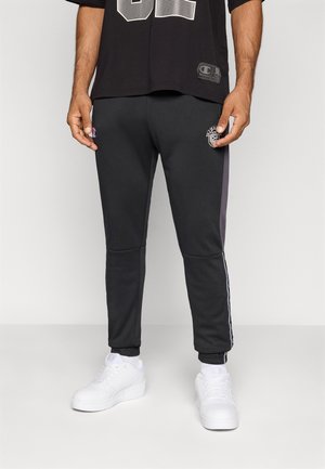 Schwarze Sweatpants mit seitlichen Streifen, die ein gesticktes Logo auf dem Oberschenkel haben. Kombiniert mit weißen Sportschuhen. Glatte Textur.