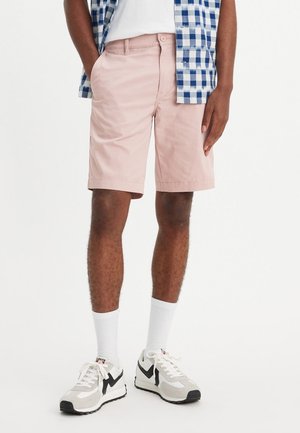 Helles rosa Shorts aus Baumwolle mit flacher Vorderseite und Seitentaschen. Kombiniert mit weißen Socken und grauen Sneakers mit schwarzen Akzenten.