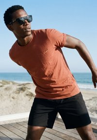 Homme portant des lunettes de soleil, un t-shirt couleur rouille et un short noir, s’appuyant de côté sur une terrasse en bois près de la plage avec l’océan en arrière-plan.
