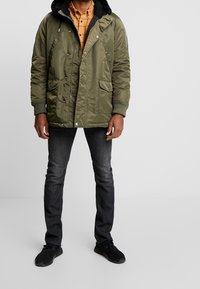 Giacca parka verde oliva con cappuccio foderato in pelliccia sintetica, tasche anteriori con bottone a scatto e polsini a coste, indossata sopra una camicia giallo a scacchi e jeans scuri.