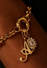 Gouden kettingarmband met een decoratieve charme met een gestileerde letter en een heldere edelsteen omringd door kleine kristallen.
