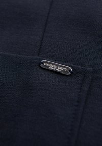 Dunkel marineblauer Stoff mit einem kleinen Metallanhänger mit der Aufschrift „OMBRE DEPT SINCE 1989“, angebracht in der Nähe einer Naht oder Taschenkante.