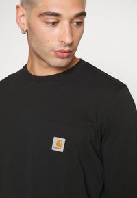 Cămașă neagră cu mânecă lungă, din bumbac, cu un buzunar pe pieptul stâng care are un eticheta pătrată ce afișează un logo galben și numele mărcii.