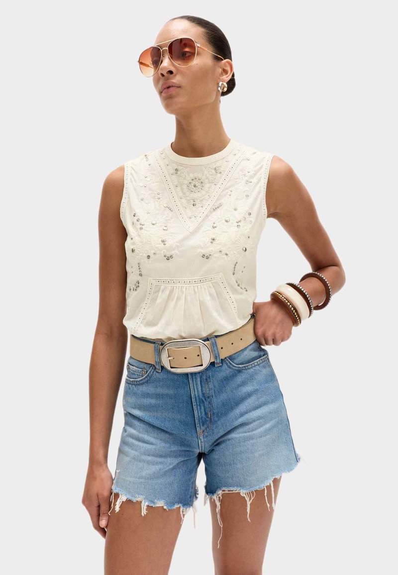 Woman wearing sleeveless white embroidered top, light blue frayed denim shorts, beige belt, sunglasses, and bracelets on right wrist.