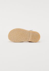 Suola di una scarpa realizzata in materiale gommoso di un marrone chiaro, con una superficie di presa modellata e una leggera curvatura nel tallone.