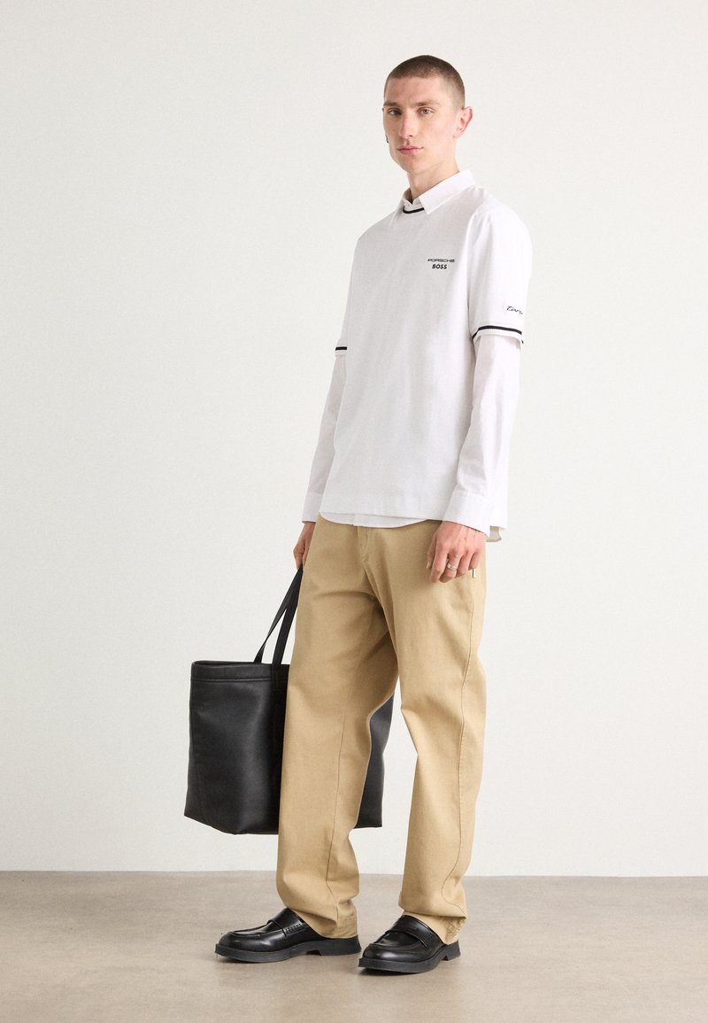 Camicia bianca a maniche lunghe con accenti neri, pantaloni beige, scarpe nere senza lacci e una borsa tote nera, su uno sfondo neutro.