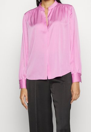 Femme portant un chemisier en soie rose à manches longues avec col bande et un pantalon noir sur mesure sur un fond uni.