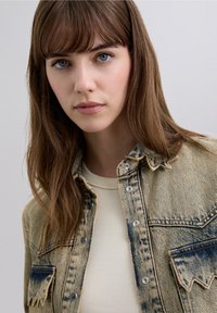 Jachetă din denim albastru decolorat cu finisaj texturat, prevăzută cu capse și două buzunare pe piept. Sub ea, se vede un top crem cu textură tip canelură.