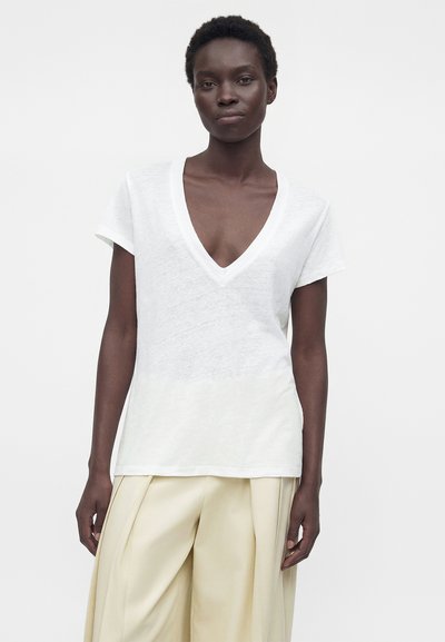 Woman with short curly hair wearing a white V-neck T-shirt and loose-fitting cream pants against a plain light background.