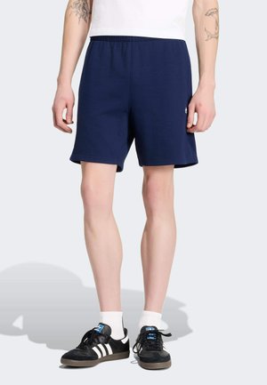 Mann trägt marineblaue Sportshorts, weiße Socken und schwarze Adidas-Sneakers, steht vor einem schlichten hellen Hintergrund.