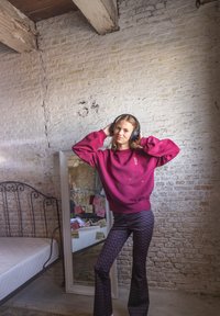 Jonge vrouw in paarse sweatshirt en met een patroonbroek past haar koptelefoon aan naast een spiegel met motiverende post-its in een rustieke kamer.