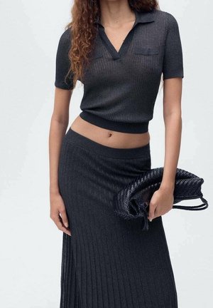 Femme portant un haut court à manches courtes en tricot côtelé foncé et une jupe taille haute assortie, tenant une pochette noire tissée.