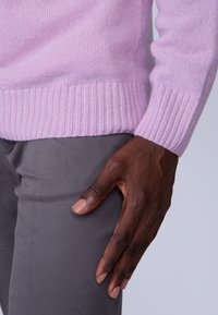 Pull en cachemire violet clair avec un ourlet côtelé, associé à un pantalon gris. Une main repose sur la cuisse, mettant en valeur le teint et la texture de la peau.