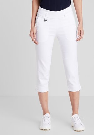 Femme portant un pantalon court blanc et des baskets blanches, debout devant un fond gris clair, les mains détendues le long du corps.
