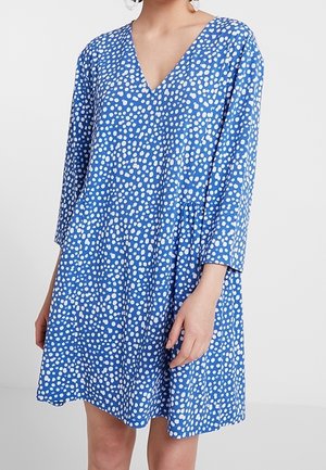 Robe bleue à col en V avec des pois blancs irréguliers, manches trois-quarts et coupe ample au-dessus du genou, portée par une personne debout.