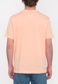 T-shirt en coton de couleur pêche clair avec un col rond et des manches courtes, affichant un ourlet droit et des coutures minimales, porté avec un jean bleu.