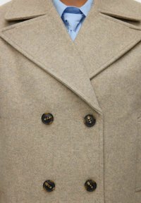 Bēšas krāsas vilnas maisījuma divrindu mētelis ar platu atloku un četriem tumšiem pogām. Apakšā gaiši zila krekla ar apkakli un saskaņotu kaklasaiti.