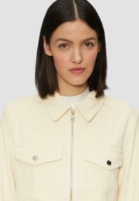 Vrouw met steil donker haar draagt een crèmekleurige ritsjack met klepzakken tegen een lichte achtergrond.