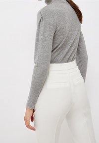 Grauer Strickrollkragenpullover mit taillierter Silhouette, kombiniert mit hochgeschnittenen weißen Hosen, die über rückwärtige Taschen und doppelte Gürtelschlaufen verfügen.