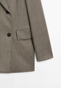 Blazer marron sur mesure avec boutons noirs et poche à rabat sur fond blanc, montrant partiellement le devant et la manche droite.
