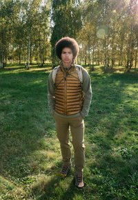 Brown puffer vest over a gray sweater, beige pants, and brown hiking boots. Outdoor setting with trees in the background.