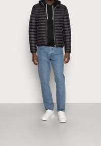 Hombre con chaqueta acolchada negra, sudadera negra con cordones blancos, jeans azul claro y zapatillas blancas de pie contra una pared blanca.