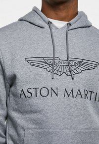 Grå huvtröja i mjukt material med dragsko i huvan. Har en svart Aston Martin-logotyp och text över bröstet. Främre känguruficka.