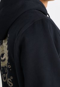 Sweat à capuche noir avec cordon de serrage, texture douce, présentant un motif graphique beige sur le devant et une coupe décontractée aux épaules.