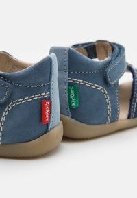 Sandalias de ante azul con una correa de velcro, suelas de goma beige, costuras blancas y etiquetas de la marca en rojo y verde en el talón.