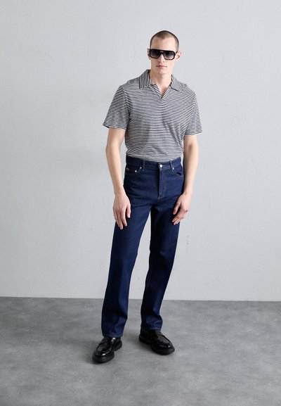 Camisa de rayas de manga corta en blanco y negro, combinada con pantalones vaqueros oscuros de cintura alta. El modelo lleva gafas de sol oscuras y zapatos negros.