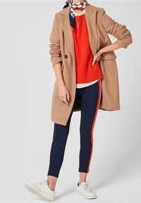 Manteau beige en mélange de laine, pull rouge, pantalon bleu marine avec des bandes rouges sur les côtés, baskets blanches et écharpe à motifs autour du cou.